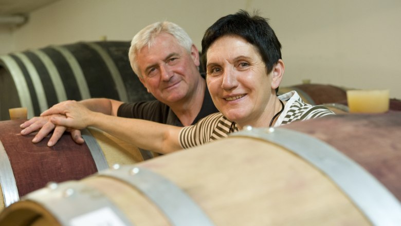 Two people smile next to wooden barrels in a winery.