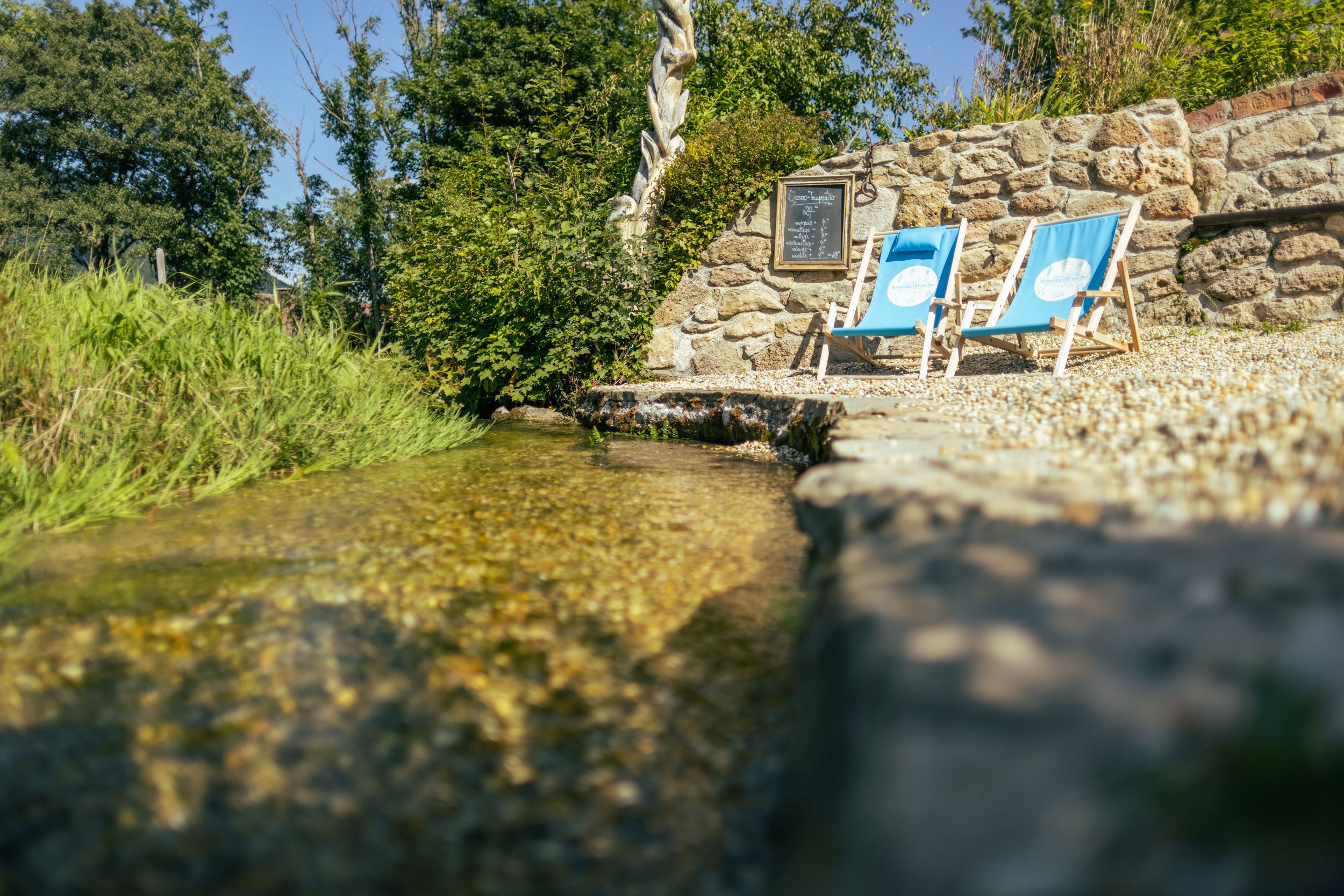 Two blue deckchairs stand next to a small stream in front of a stone wall.