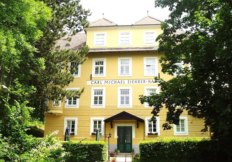 Front view of the Carl Michael Ziehrer House, a yellow building with several windows and a green entrance, surrounded by trees.