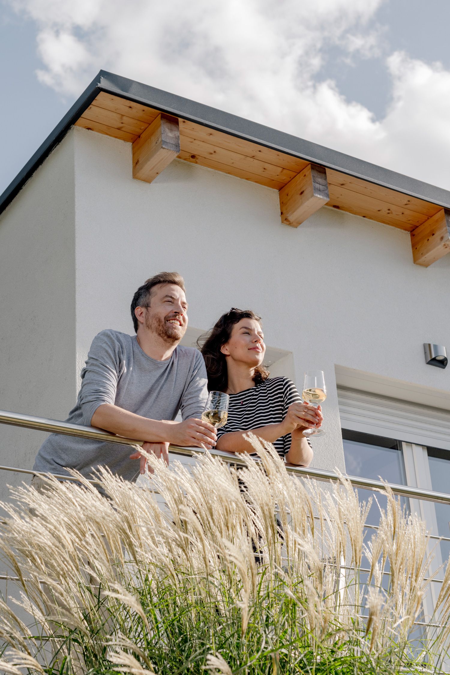 A couple stands on a balcony holding glasses of wine, with a modern building in the background.
