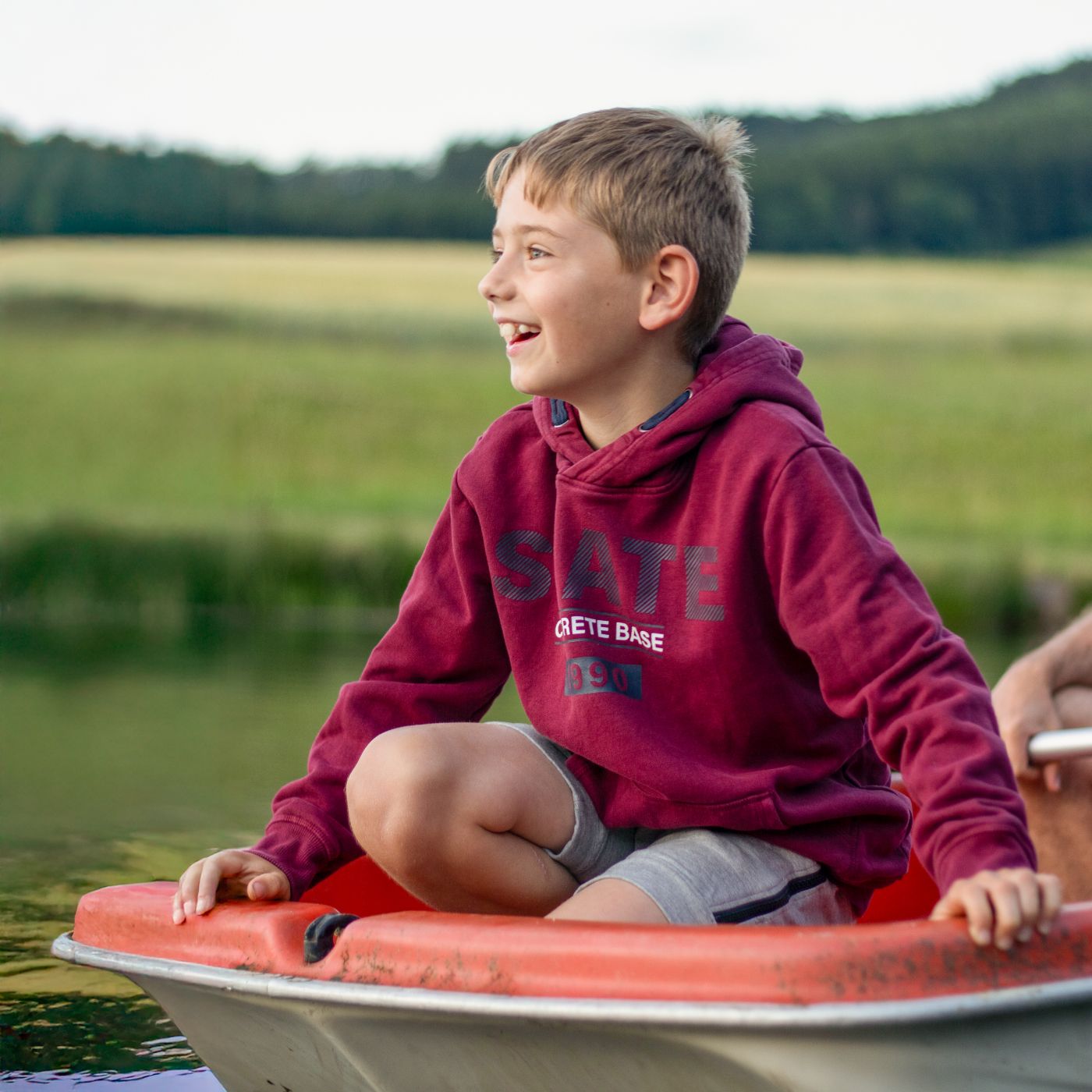 A boy in a red hoodie sits smiling on a boat on a pond.
