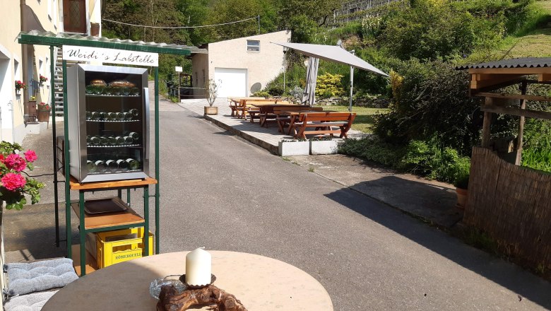 An outdoor area with a drinks fridge, tables and benches next to a vineyard.
