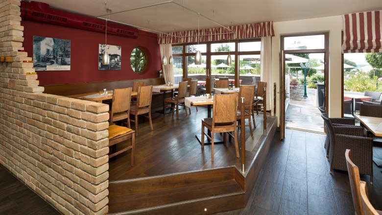 Ein gemütliches Restaurant mit Holzmöbeln, roten Wänden und großen Fenstern, die auf eine Terrasse führen.