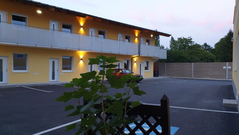 Exterior view, © Alexandra Heiling-Niegl Exterior view of a two-story building with yellow facade and illuminated balcony, in front of it a parked red car and plants in the foreground.