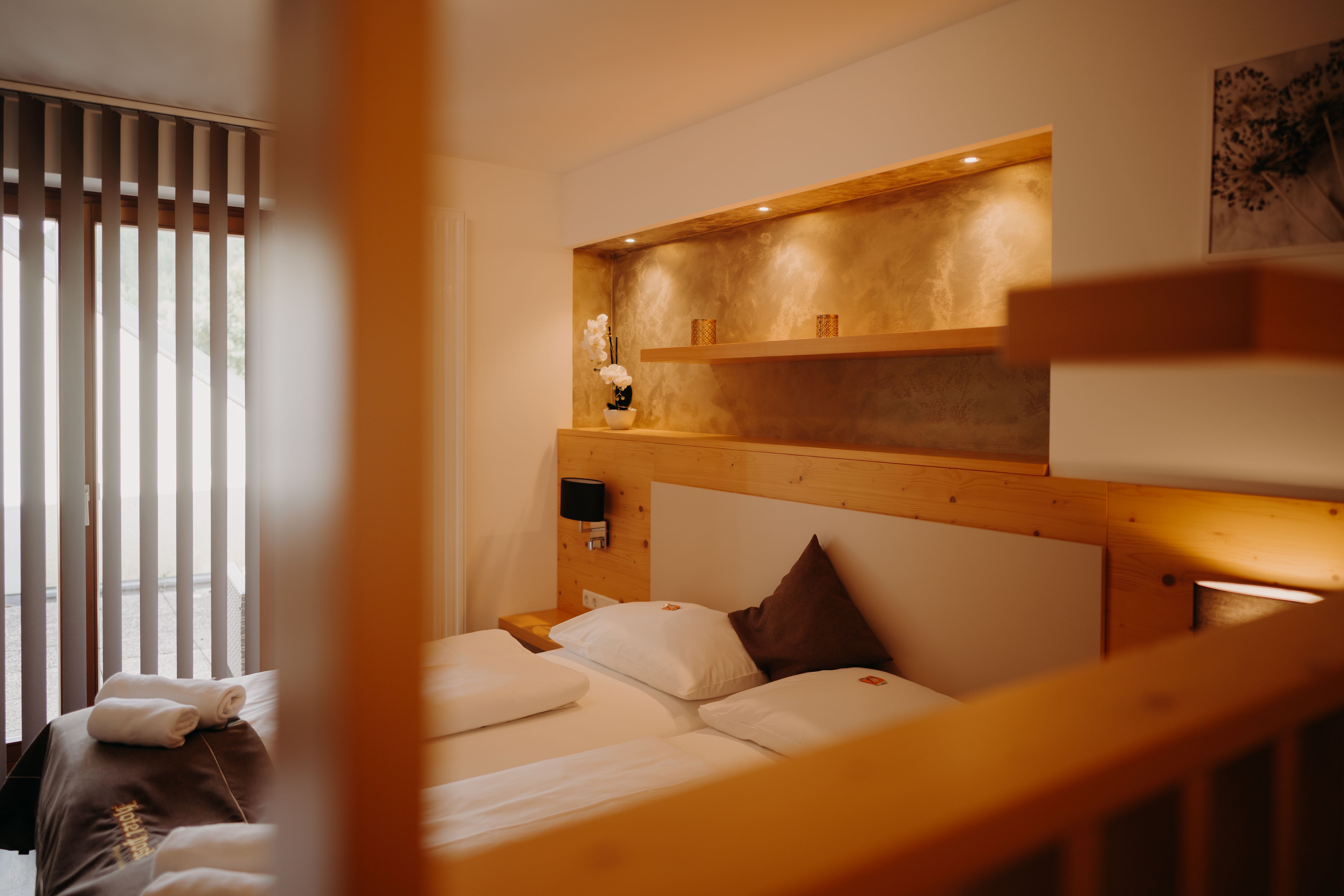 A cozy apartment with a double bed and a balcony door. Part of the wall is paneled with light-colored wood; behind the bed is a recessed niche with a shelf and spotlights, painted in an earthy gold.