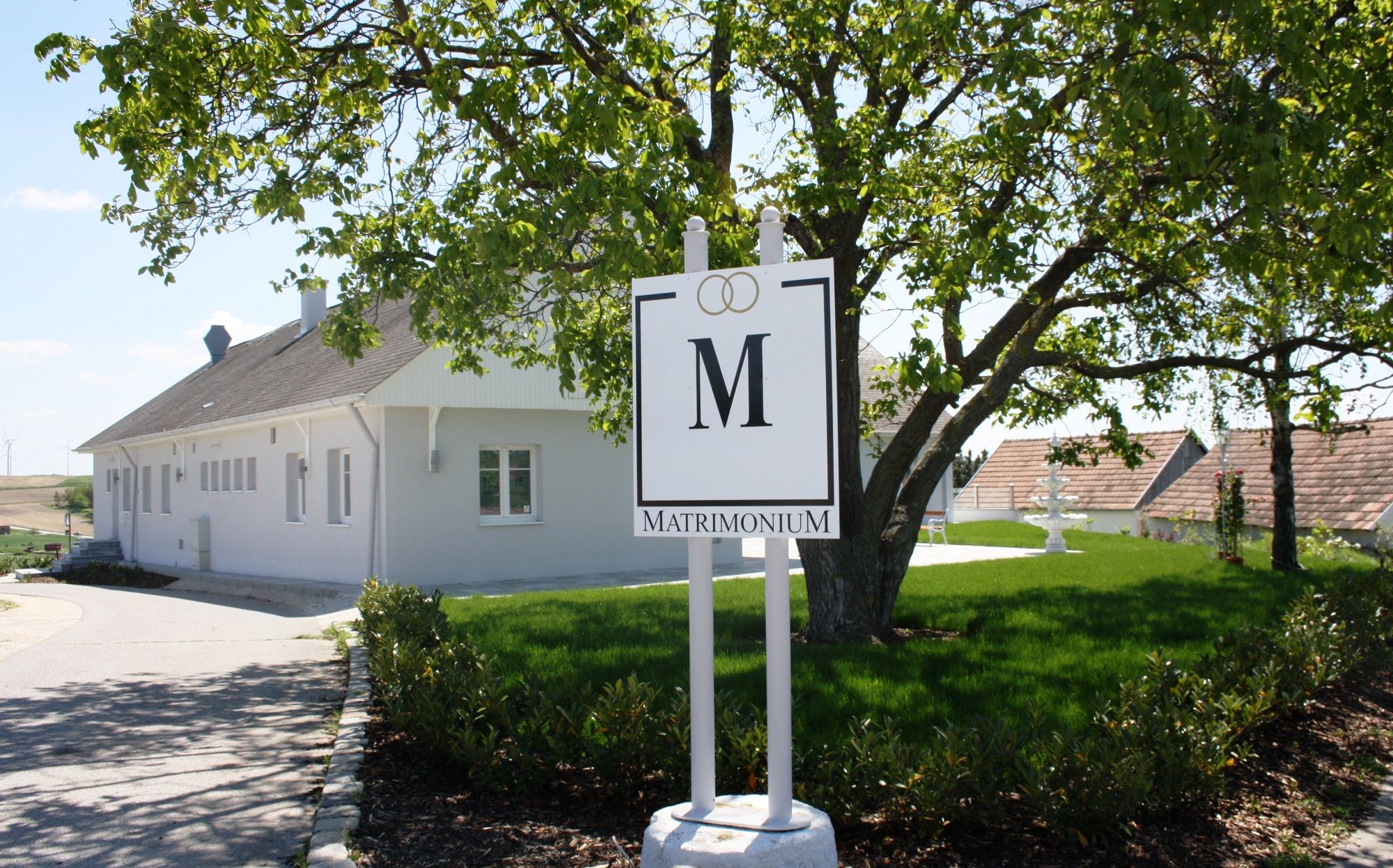 A white building with a "Matrimonium" sign in the foreground, surrounded by a green landscape.