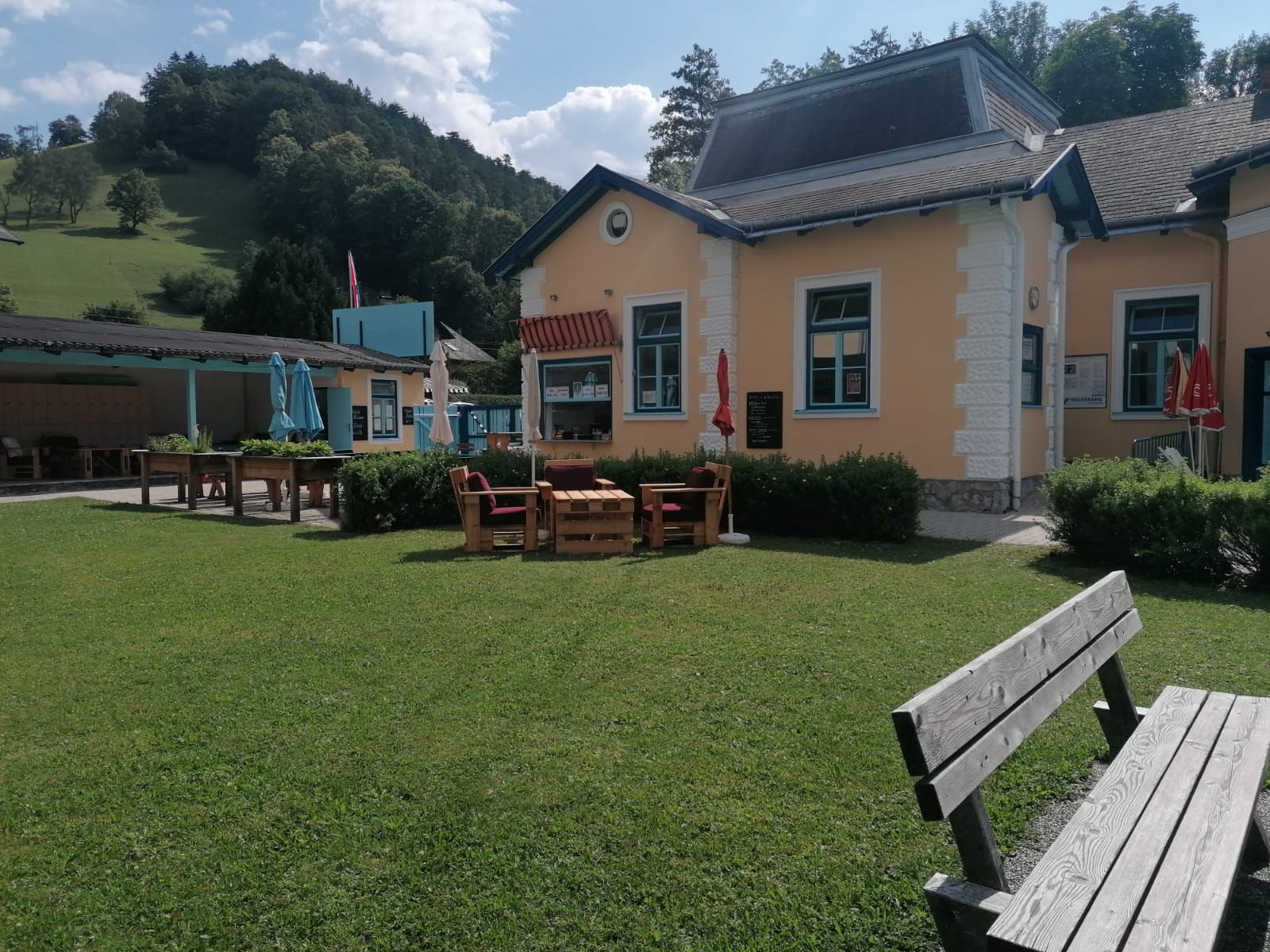 A yellow building with blue window frames, surrounded by a green meadow and benches, in front of a wooded hill.