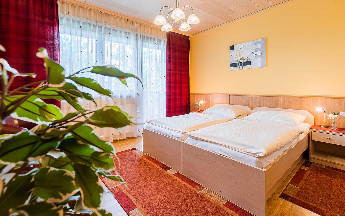 Cozy bedroom with double bed, yellow wall, red carpet and plants in the foreground.