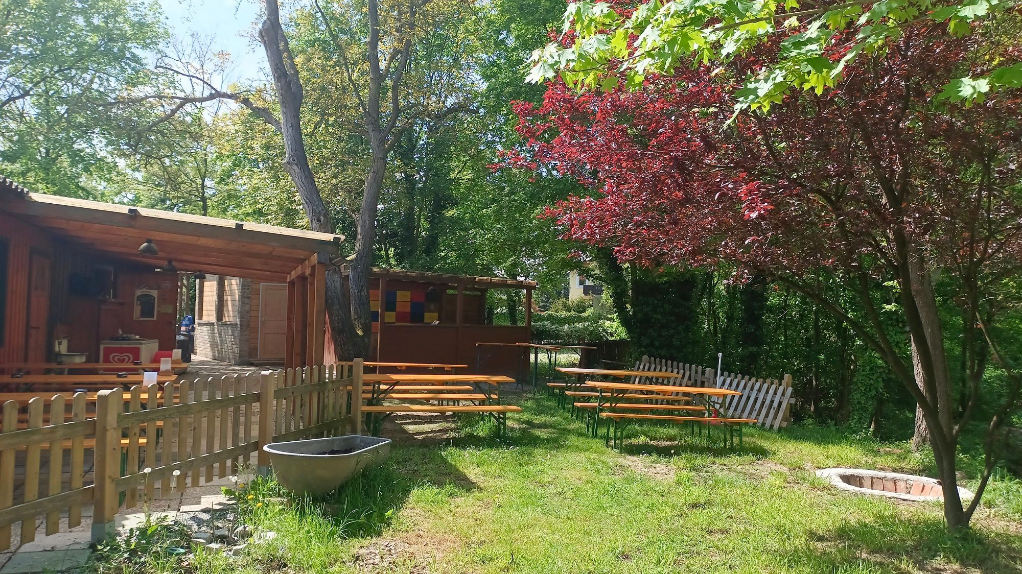 A beer garden with wooden benches and outdoor tables, surrounded by trees and a wooden fence.