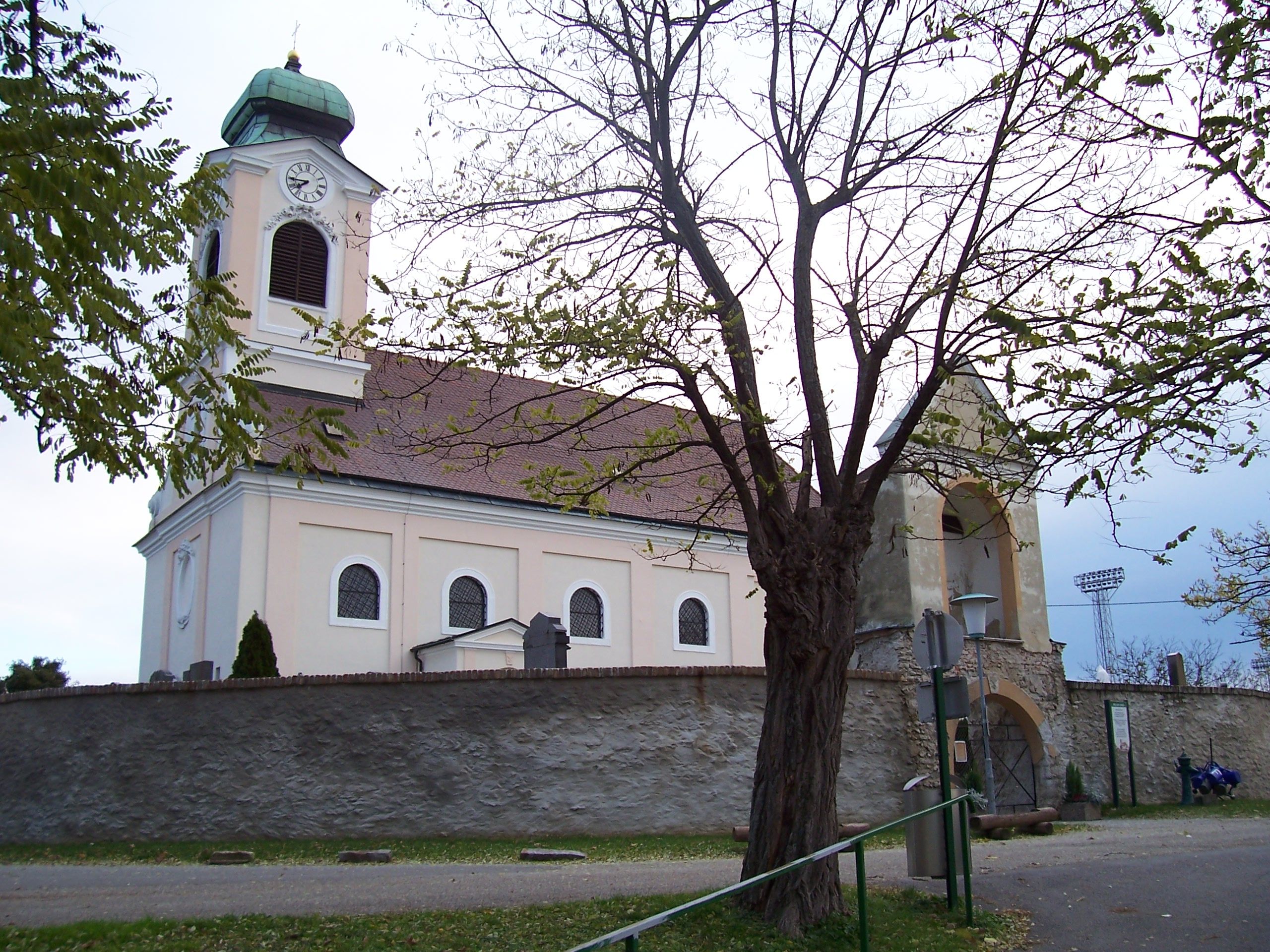 Church with tower and clock, surrounded by trees and a stone wall.