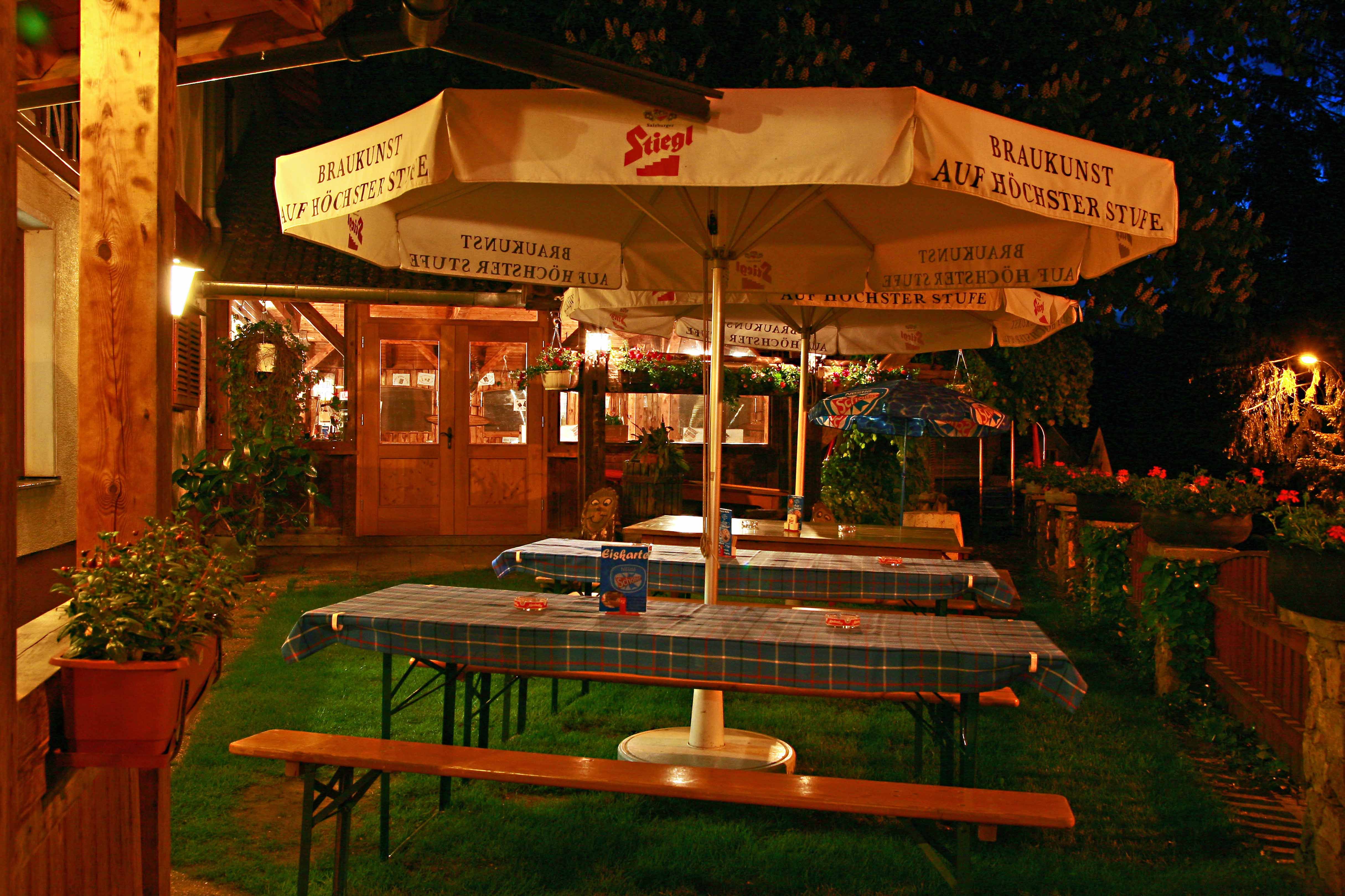 A cozy beer garden at night with wooden tables, blue checkered tablecloths and large Stiegl umbrellas.