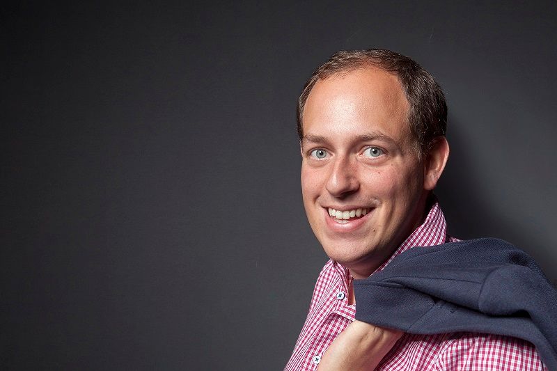 Man in plaid shirt with jacket over his shoulder in front of a gray background.