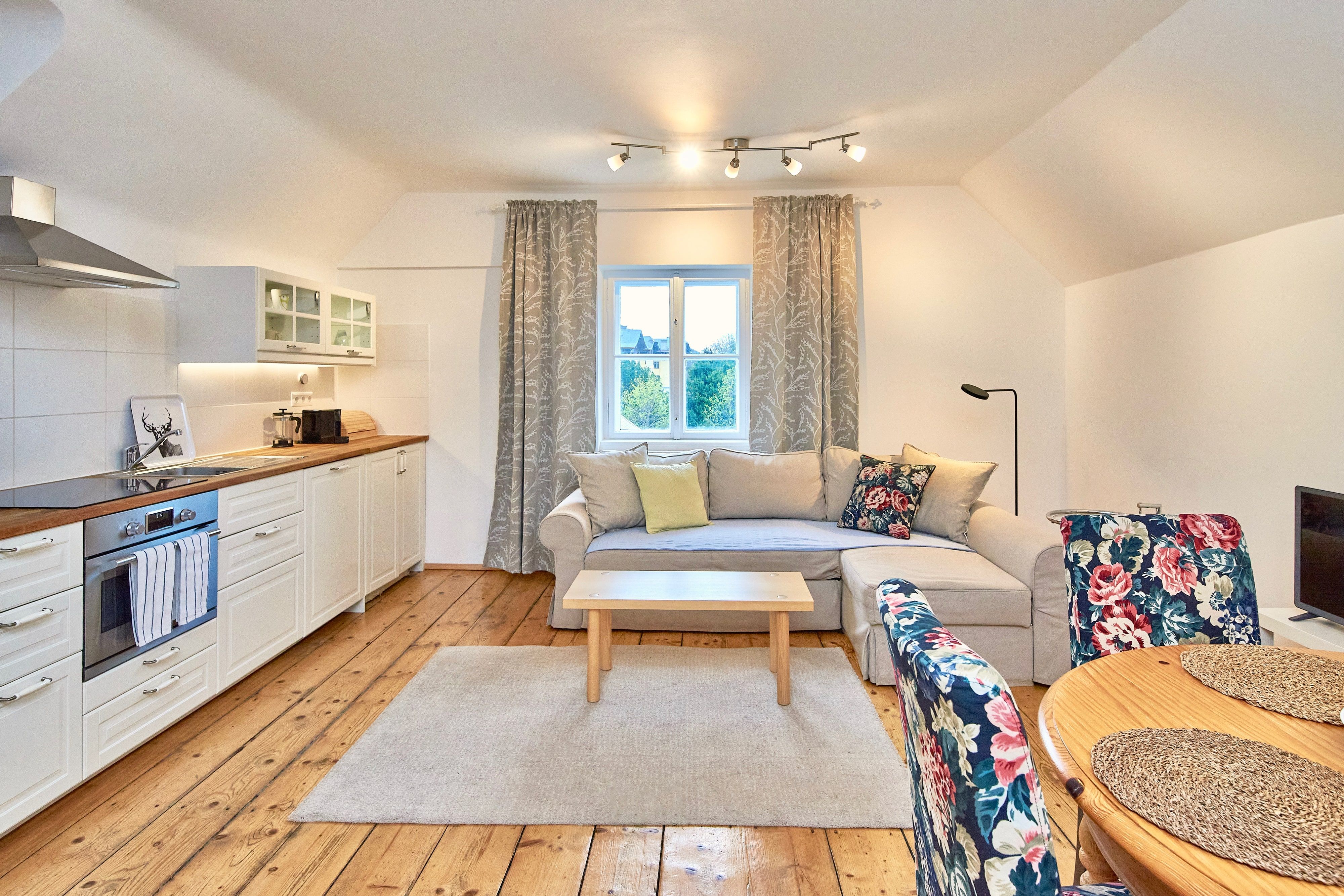 Cozy living room with open kitchen, wooden floor, beige couch, floral patterned chairs and a window with curtains.