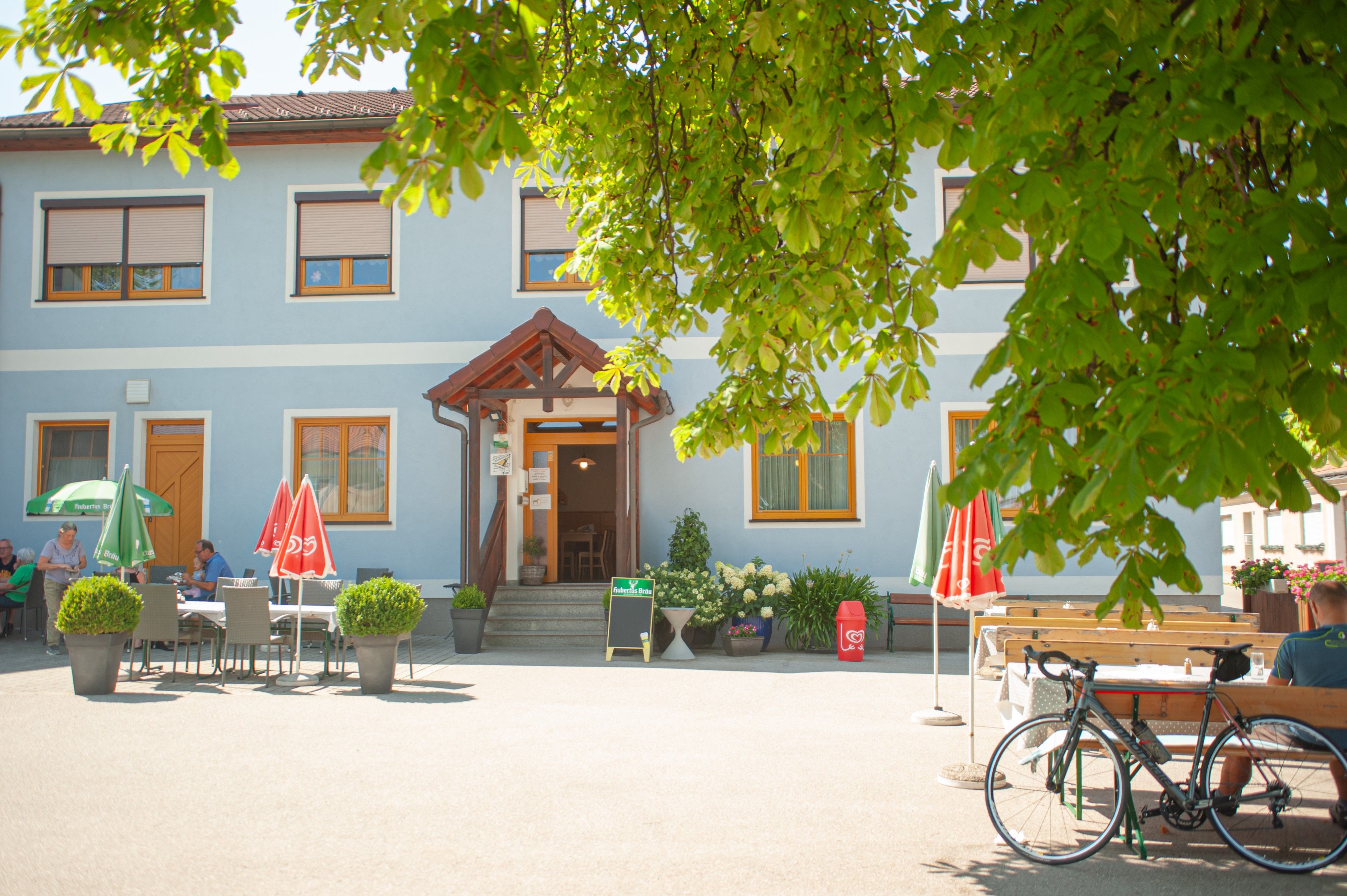 A blue inn with parasols and tables outside, surrounded by green trees.