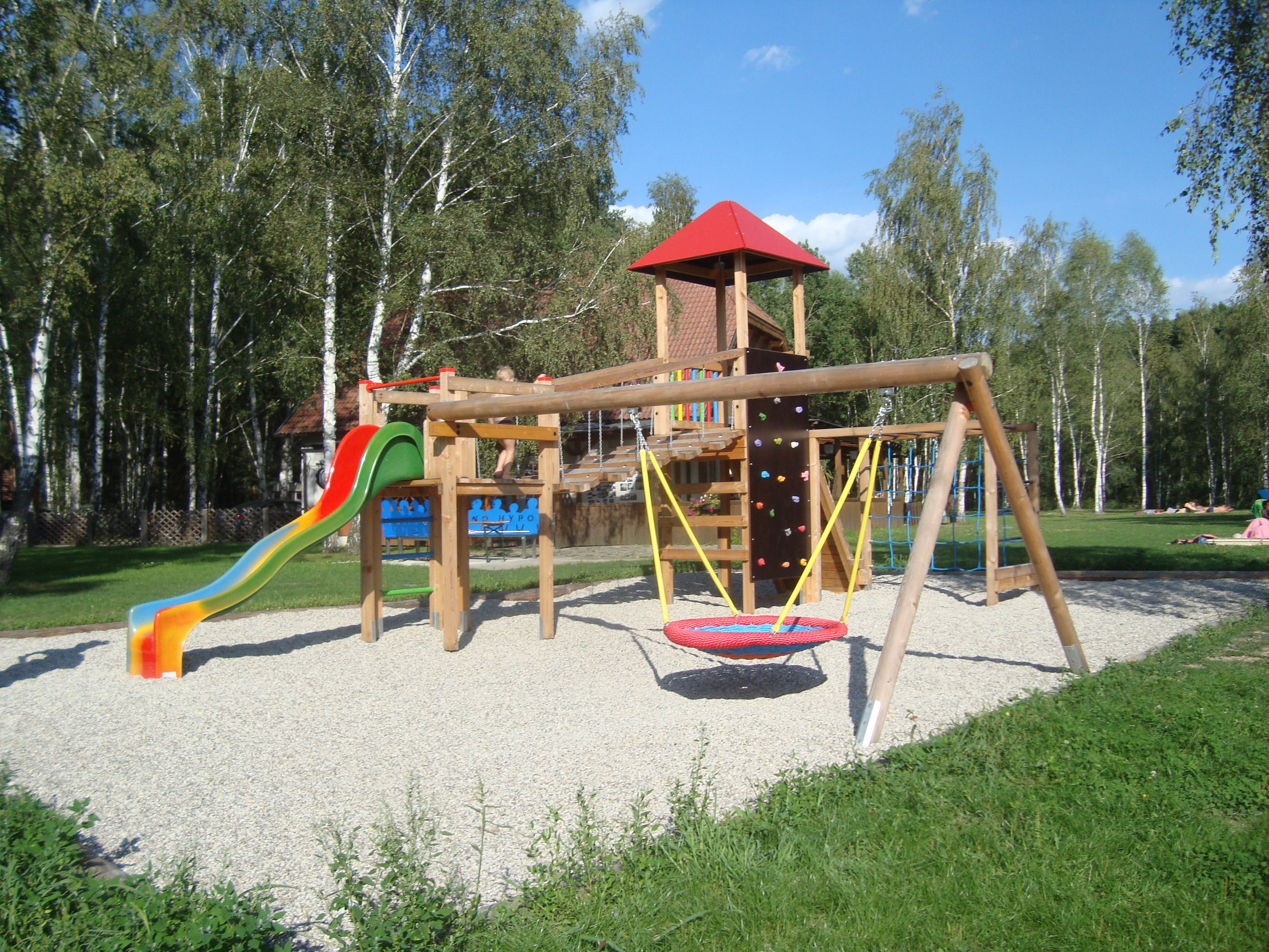 A children's playground with slide, swings and climbing frame, surrounded by trees.