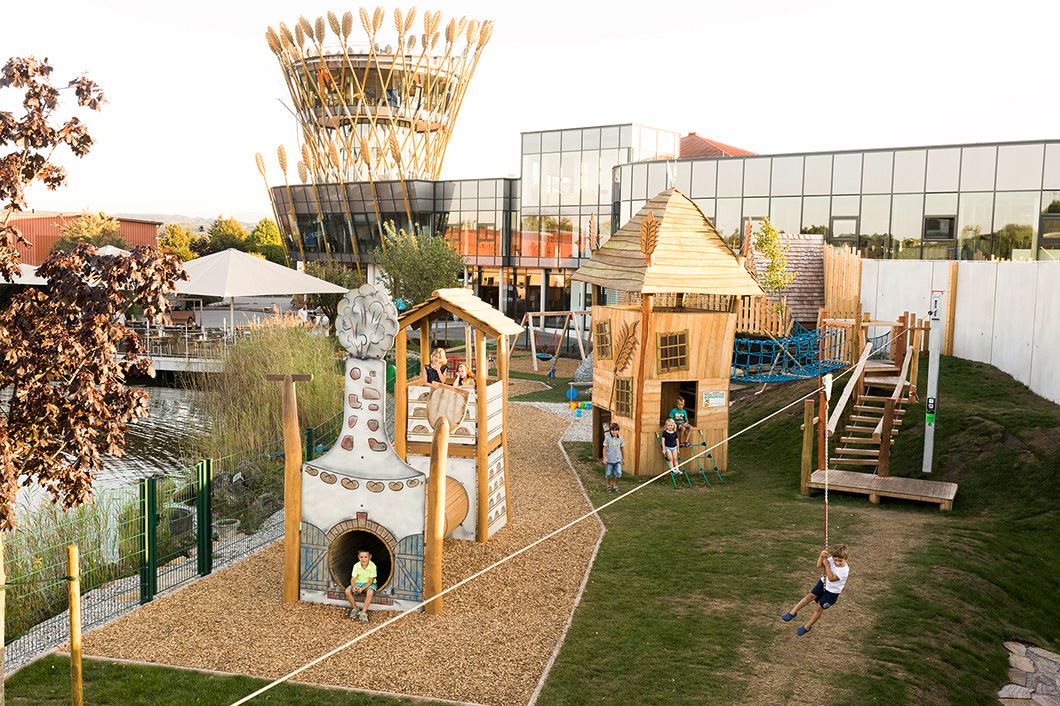 A modern playground with wooden houses and a rope slide in front of a building with a glass façade and a tower with wheat ornaments.