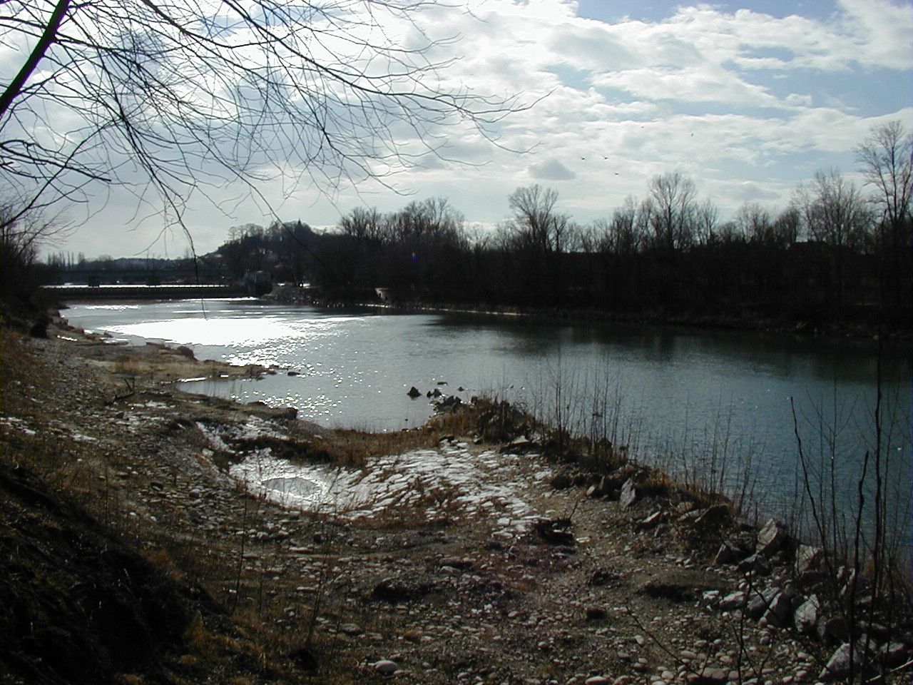 A calm river with bare trees on the banks under a cloudy sky.