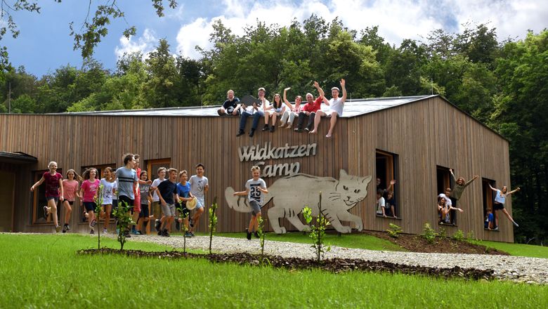A group of children and adults are sitting on a wooden building labeled 'Wildcat Camp'. Some children are standing and jumping in front of it.