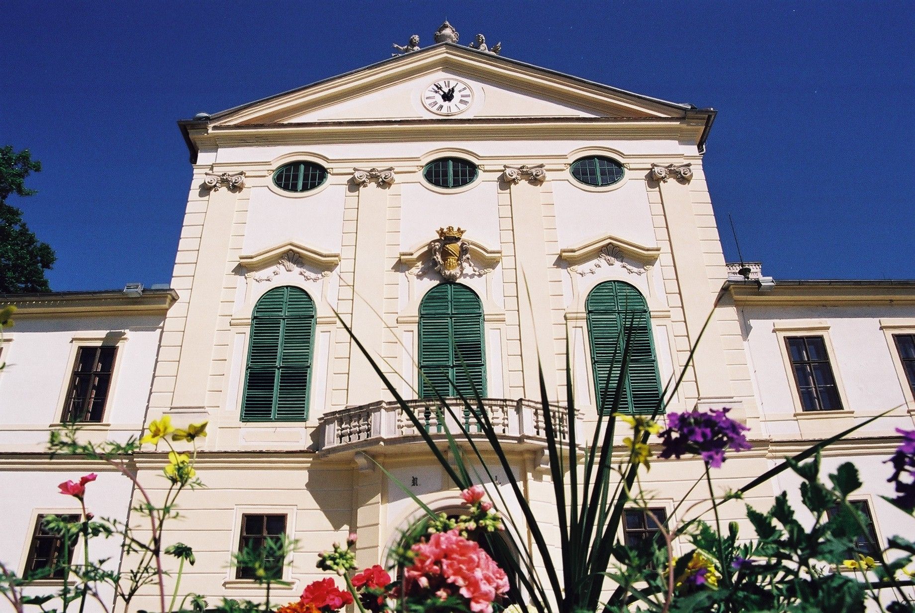 Facade of Kirchstetten Castle with green shutters and clock, surrounded by colorful flowers.