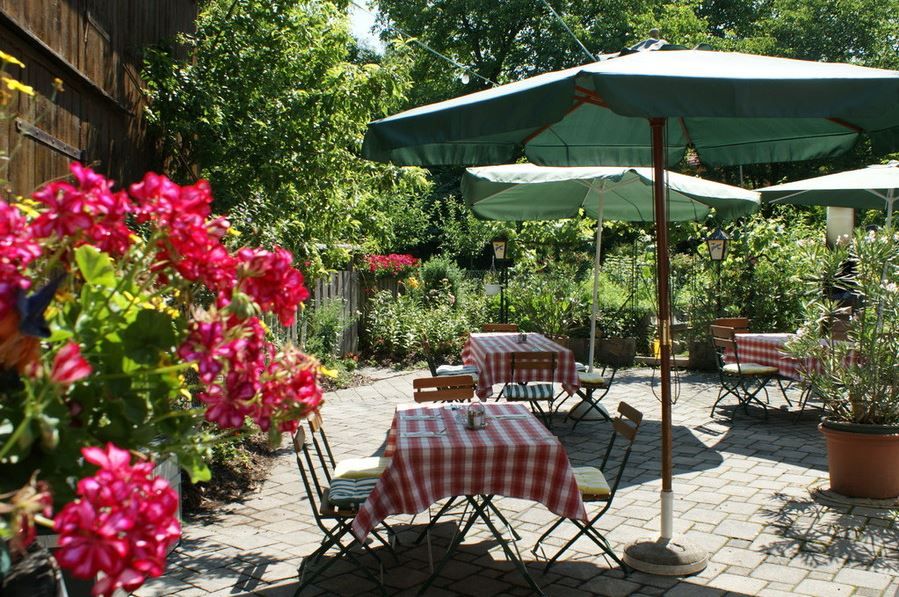 A cozy guest garden with red and white checkered tablecloths, green parasols and blooming flowers in the foreground.