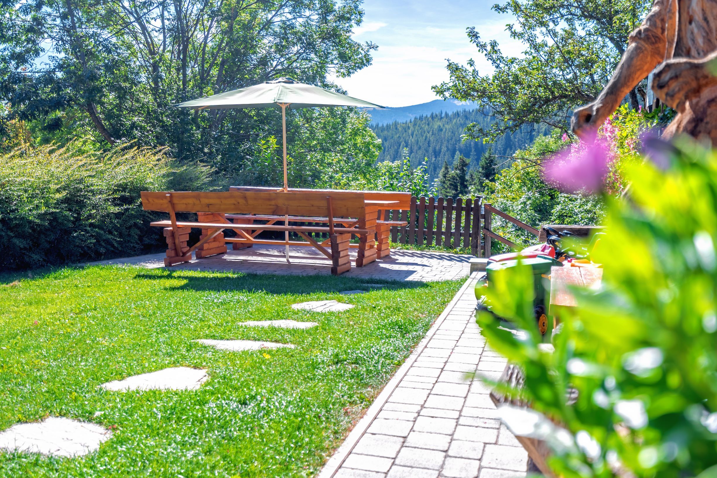 Garden with wooden table, benches and parasol, surrounded by trees and meadow.