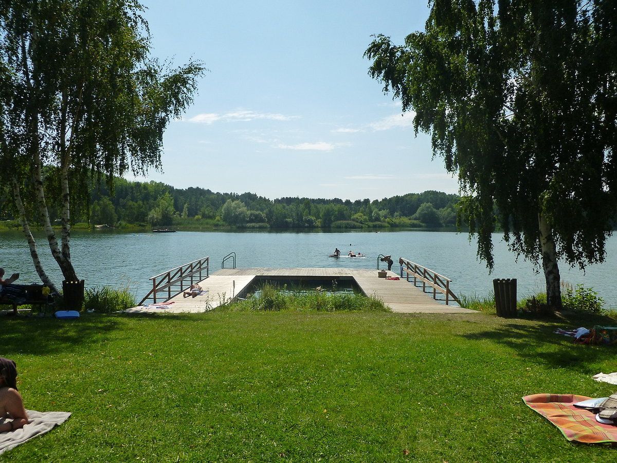 A jetty at Bergwerksee Langau with people swimming and sunbathing. Surrounded by trees and green meadows.