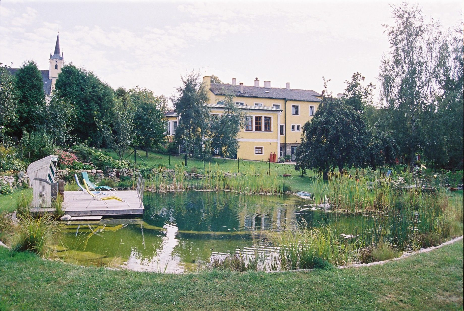 A swimming pool in a large garden with a yellow building in the background.