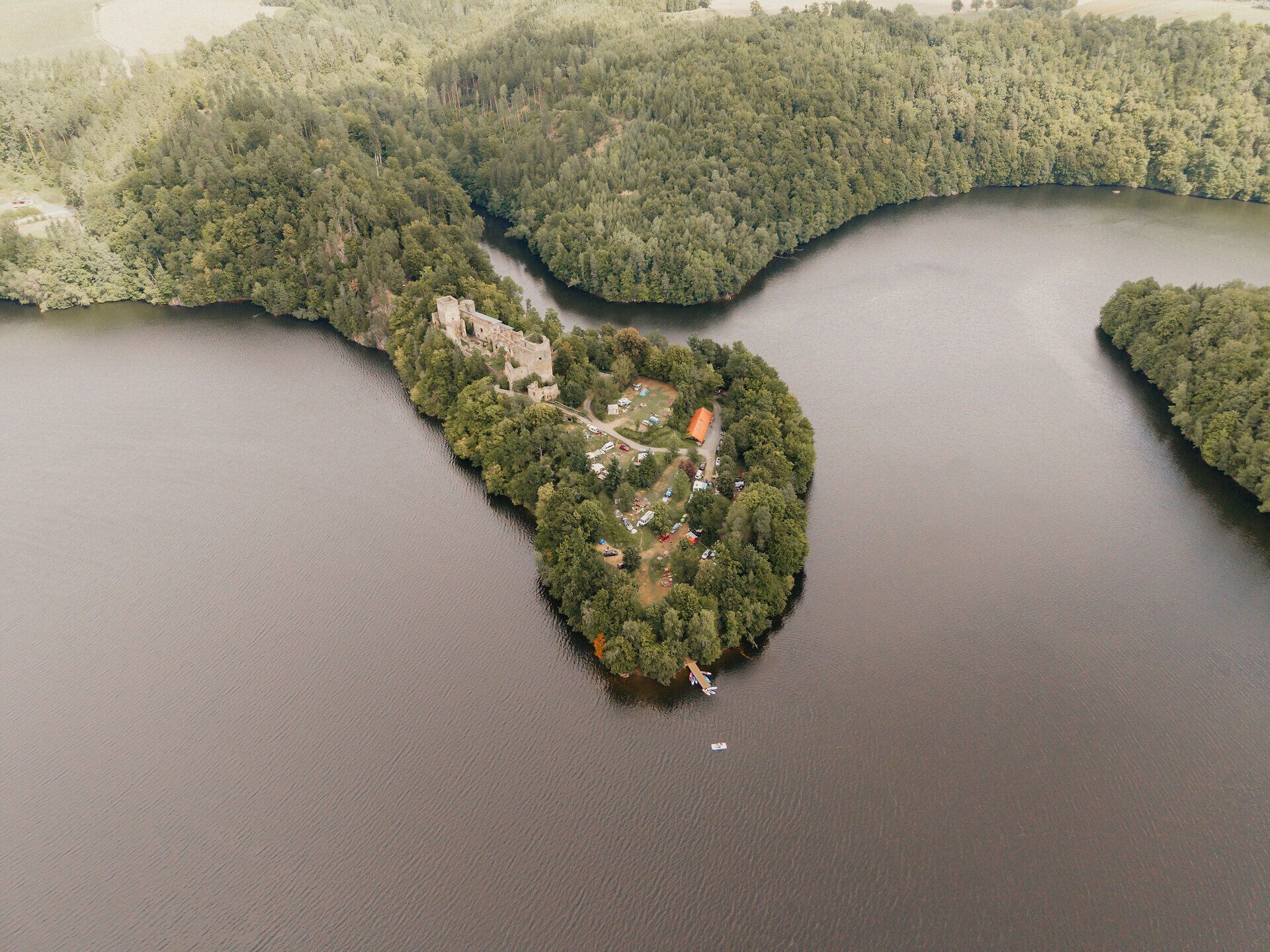 Drone shot of the wooded peninsula with the Dobra ruins in the Dobra reservoir; the ruins are surrounded by water, with small boats and meadow areas on the shore.