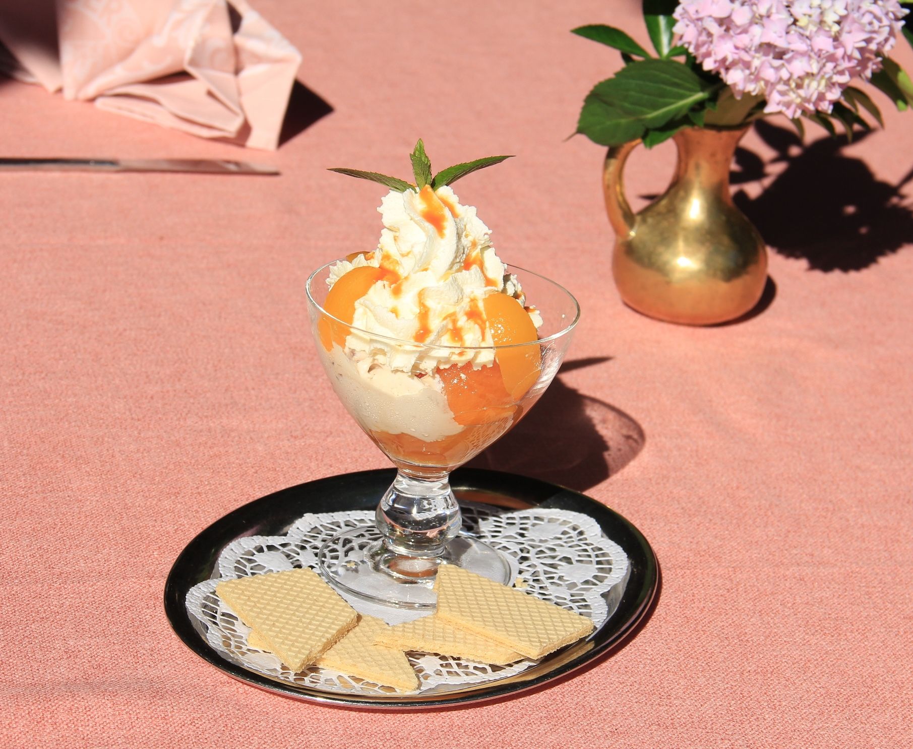 A dessert glass with ice cream, whipped cream and apricots on a tray with waffles, next to a vase with flowers.