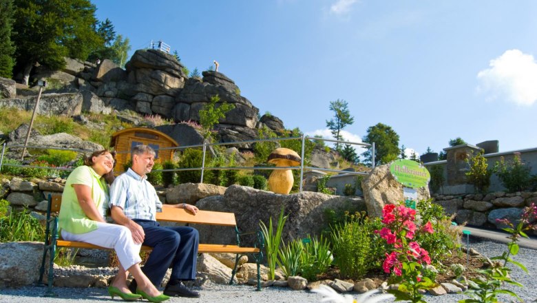 A couple sits on a bench in a herb garden with rocks in the background.