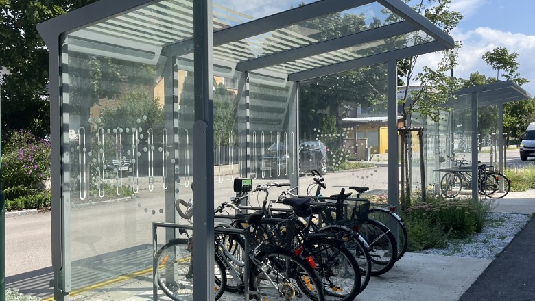 Covered e-bike charging stations with several bicycles, surrounded by trees and plants.