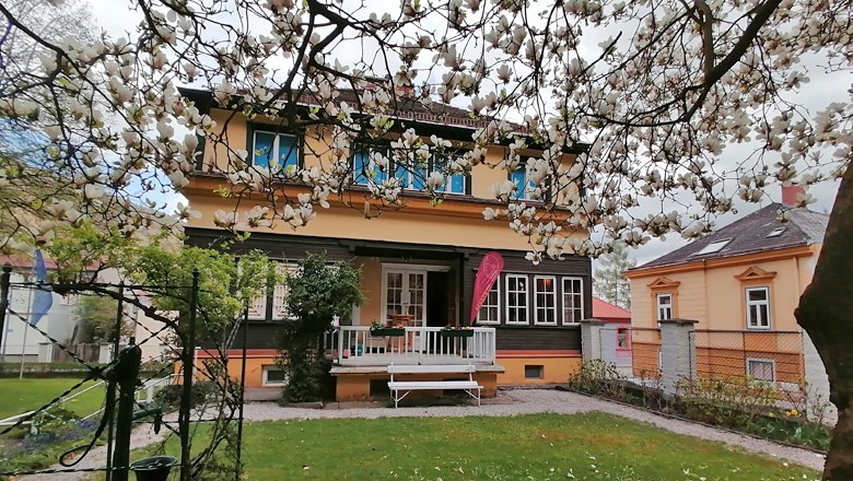 A two-story house with a yellow facade and white windows, surrounded by blooming trees.