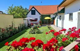 A well-tended garden with red flowers, an old plow and a house with red roof tiles in the background.