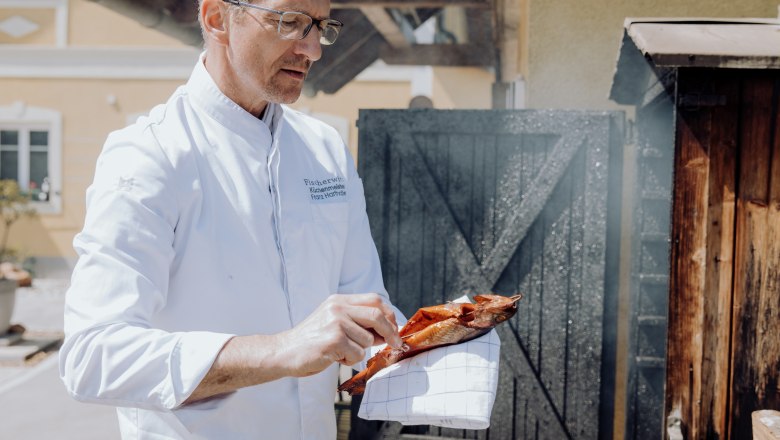 A man in a white coat holds a smoked trout in front of a smoker.