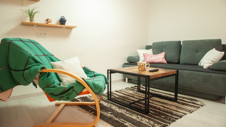 Cozy living room with grey sofa, wooden table, green rocking chair and shelves with plants.
