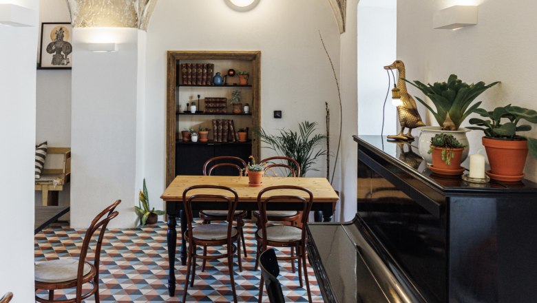 A stylish café with a vaulted ceiling, colorful tiled floor, wooden table and chairs. A piano is on the right, plants and decorations are scattered around.