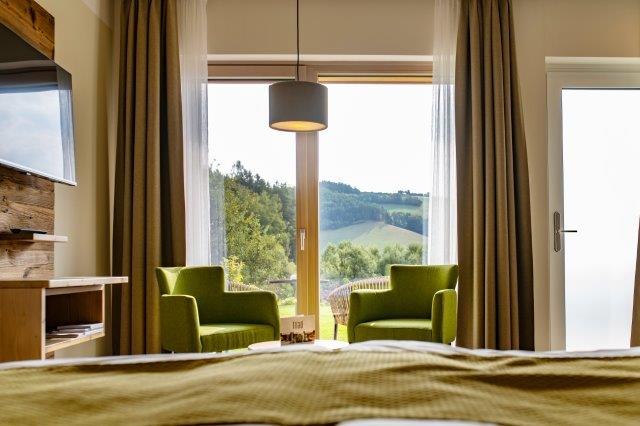 Interior view of a guest room with two green armchairs in front of a large window overlooking a hilly landscape.