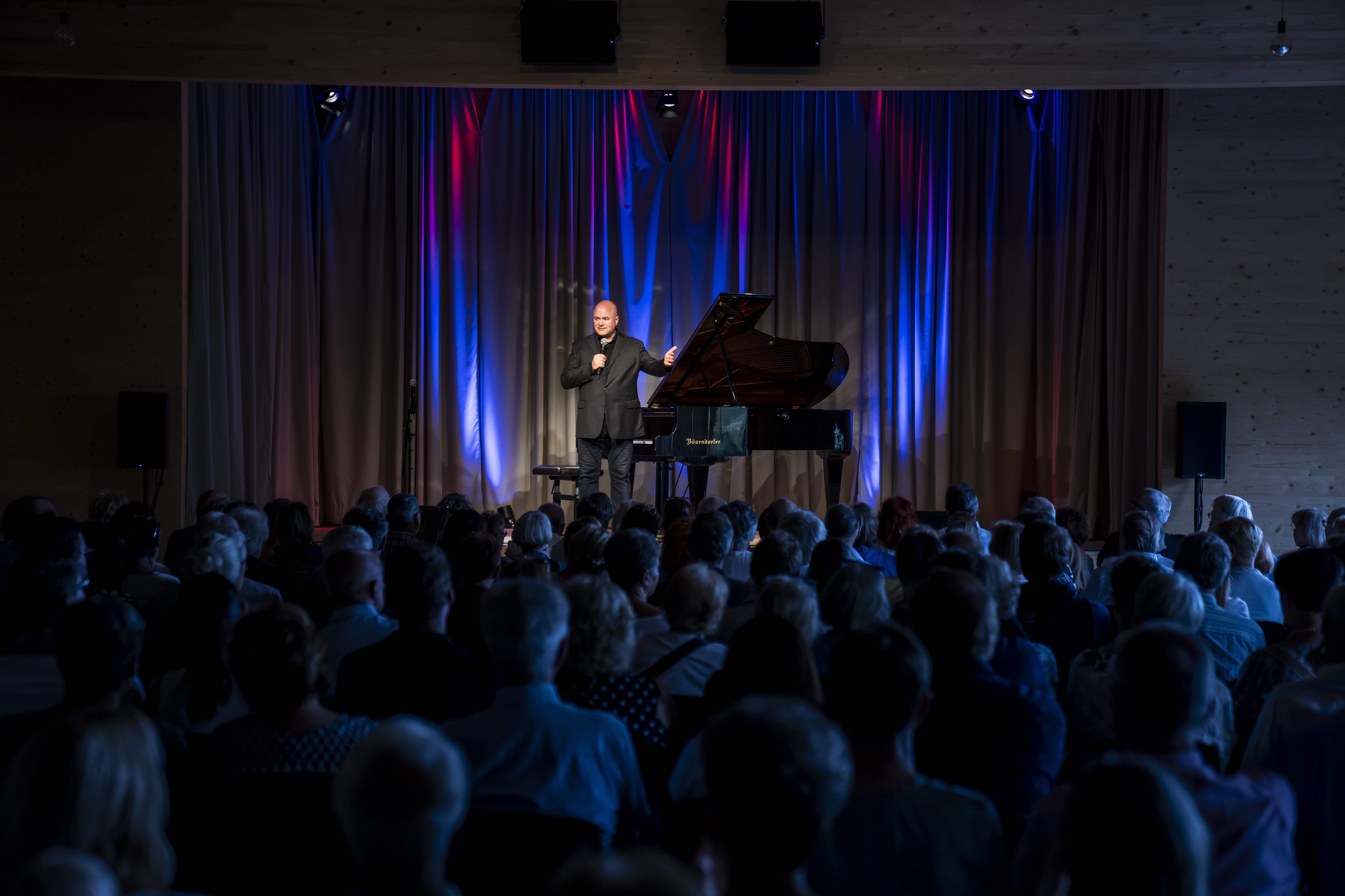 A man stands on a stage with a grand piano and speaks into a microphone in front of an audience.