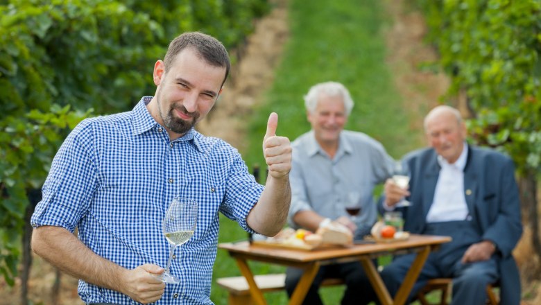 A man in a checked shirt gives a thumbs-up and holds a wine glass, while two older men sit at a table in the vineyard in the background.