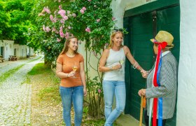 Three people are standing in front of a green gate, two are holding wine glasses, one is wearing a straw hat with colorful ribbons.