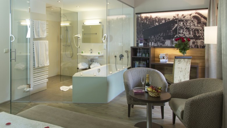 Luxurious hotel room with glass bath, cozy armchairs, table with champagne and fruit, and a large bed with rose petals.