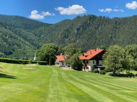 A clubhouse with a red roof in a green, mountainous landscape under a blue sky.