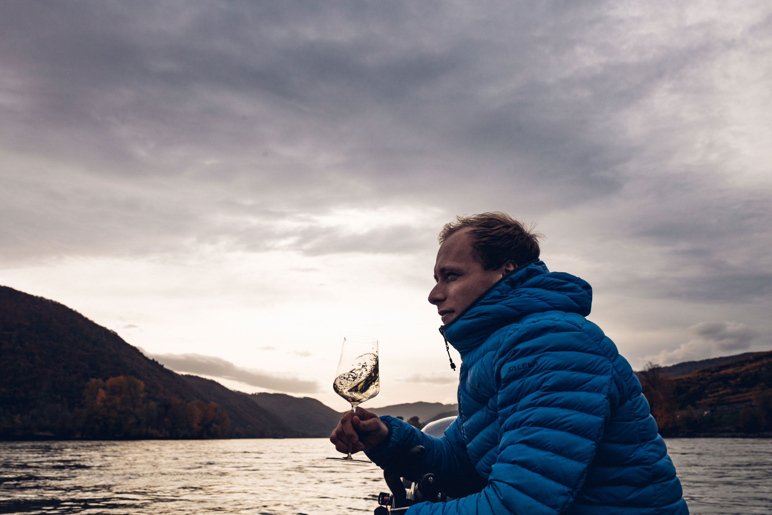 Person in blue jacket holding a wine glass on a boat in front of a river landscape.