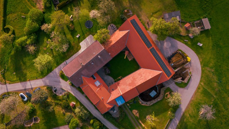 Aerial view of a farm with red roofs and solar panels, surrounded by green meadows and trees.