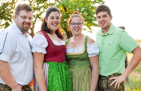 Four people in traditional costume stand smiling outdoors on a green meadow, with a tree in the background.