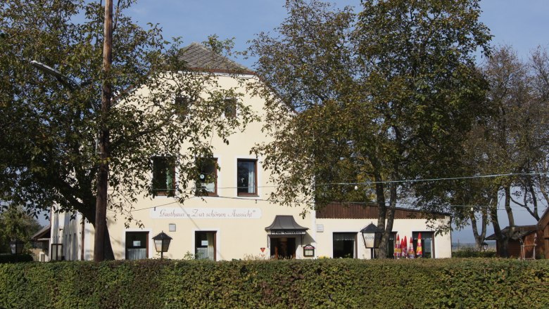 A yellow inn with the inscription 'Gasthaus Zur schönen Aussicht', surrounded by trees and hedges.