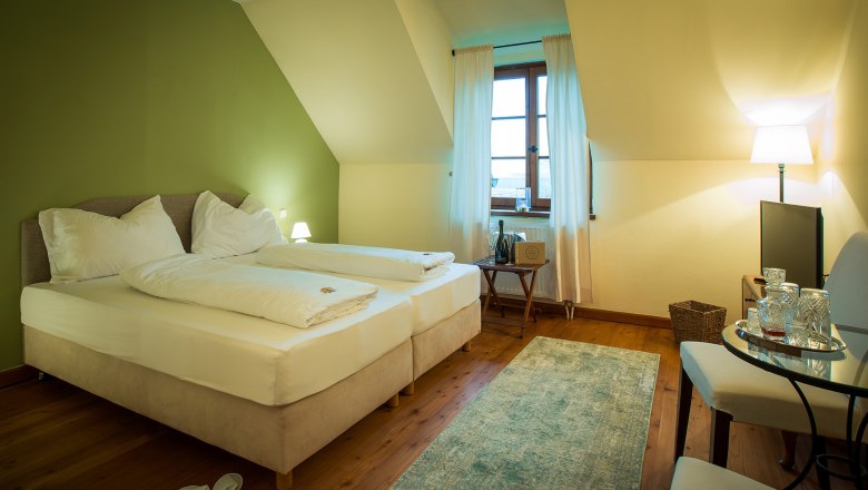 A cozy double room with a green-painted wall, two beds, a window with curtains, a small table and a TV.