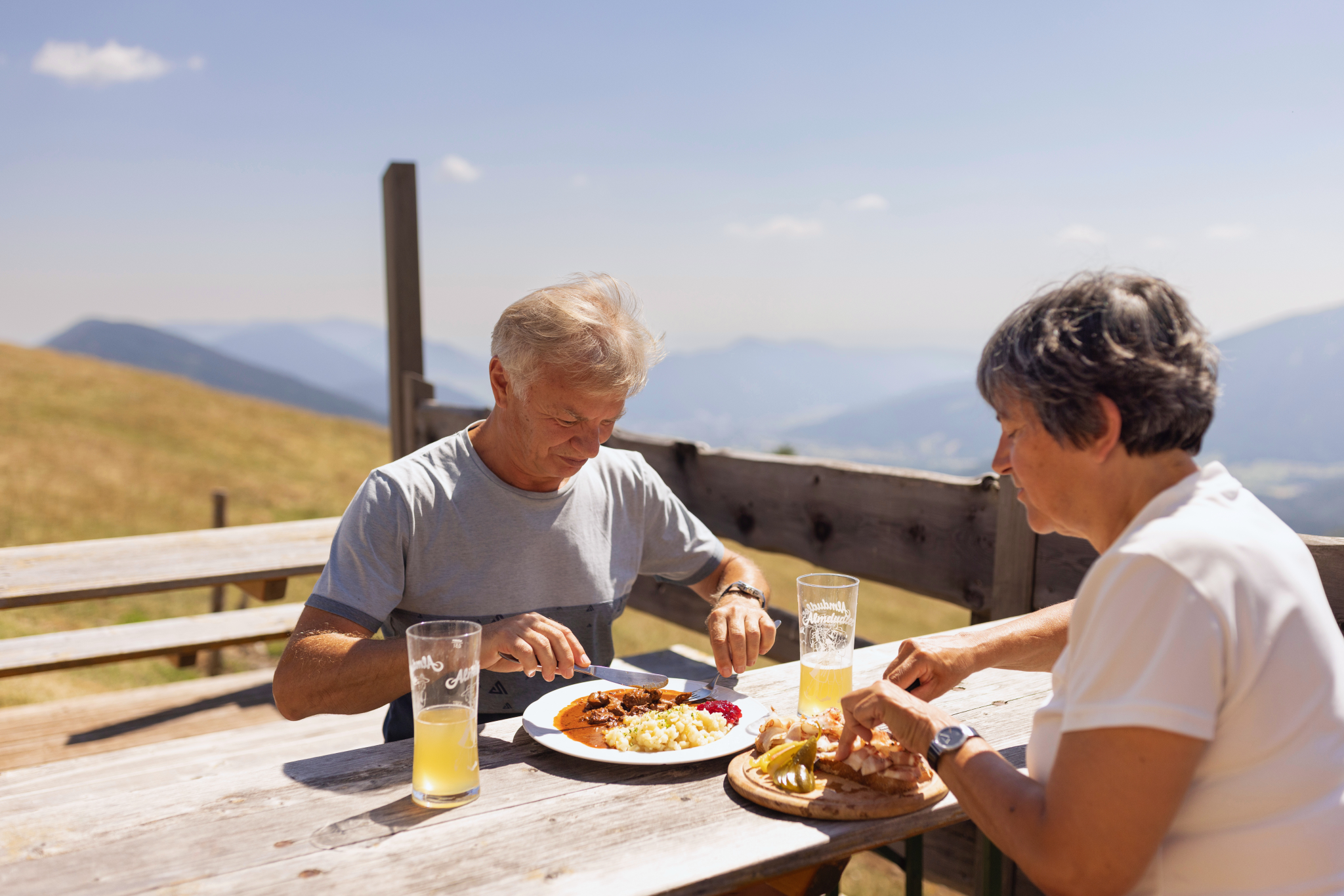 A couple eats at a wooden table outside with a view of the mountains.