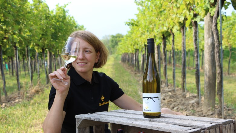 A woman in the vineyard holds a glass of white wine and sits next to a bottle of wine on a wooden crate.