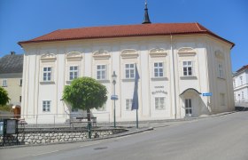 Historic building with a red roof and white walls, surrounded by trees and a road.